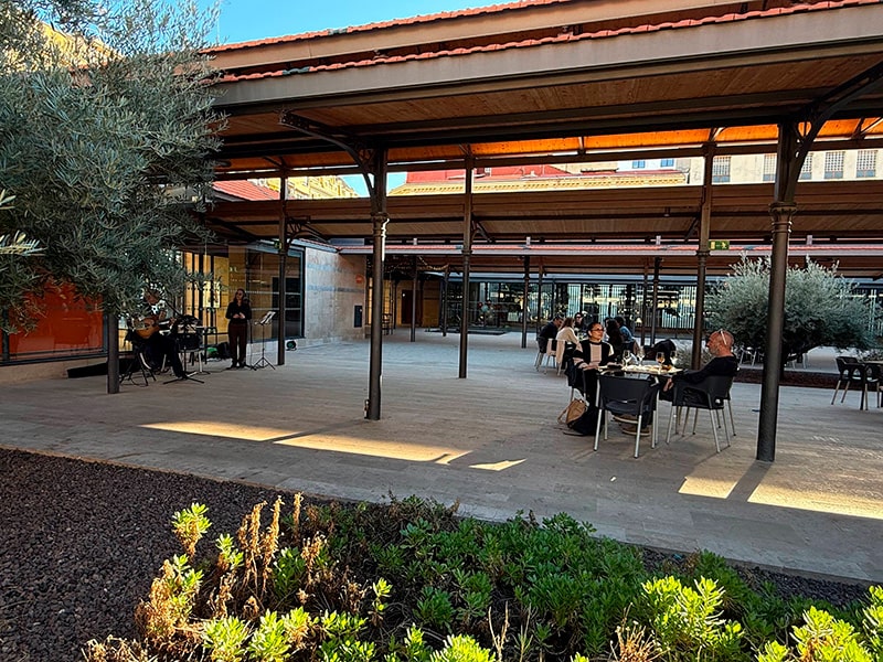 Открытый двор у Mercado del Grao с навесами, зелёными клумбами, столиками и уличными музыкантами (Кабаньяль, Валенсия).