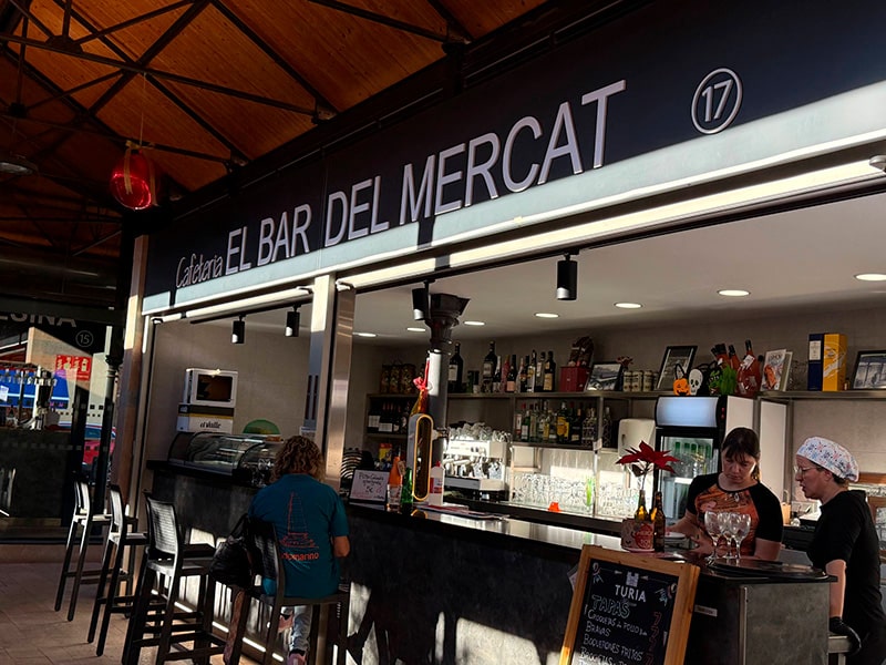 Барная стойка El Bar del Mercat внутри рынка: вывеска, напитки на полках и посетители у стойки (Кабаньял, Валенсия).