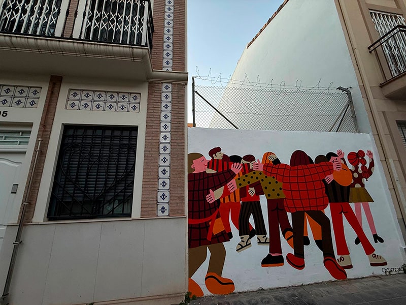 Уличное граффити с группой людей, обнимающихся и танцующих, на стене между домами в районе Кабаньял (Валенсия).