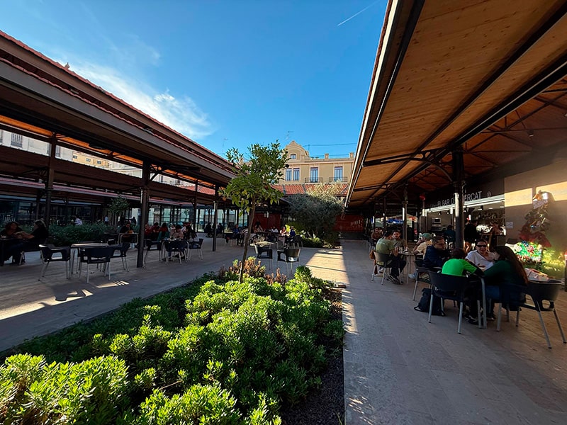 Открытая зона рынка с навесами, кафе-столиками и зелёными клумбами у Mercado del Grao в районе Кабаньял (Валенсия).