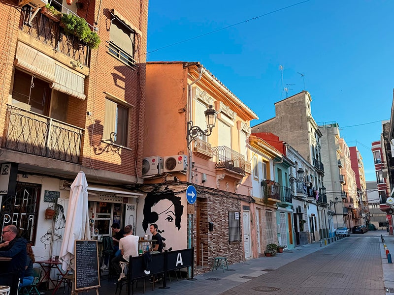 Улица в Кабаньяле с низкими разноцветными домами и уличным кафе; на стене &mdash; граффити с женским портретом (Валенсия).