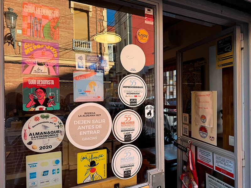 Витрина ресторана La Aldeana с наклейками наград, рекомендациями гидов и афишами на стекле в районе Кабаньял (Валенсия).