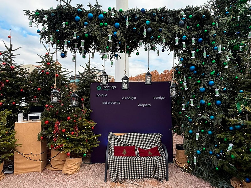 Рождественская фотозона на Plaza de Colón в Мадриде: арка из еловых веток с гирляндами и шарами, вокруг — украшенные елки и лавочка для фото.