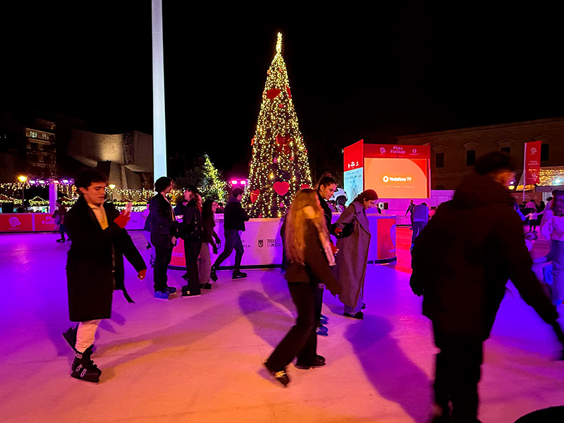 Каток на Plaza de Colón в Мадриде вечером: люди катаются на коньках рядом с большой рождественской ёлкой и праздничной подсветкой