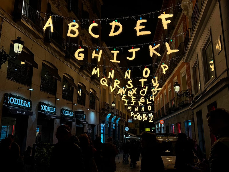 Световая рождественская иллюминация на Plaza del Callao: над улицей подвешены светящиеся буквы алфавита, внизу — вечерняя толпа и витрины магазинов.