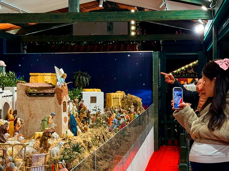 Посетители рождественского рынка фотографируют большой вертеп: женщина снимает на телефон, мужчина показывает на фигуры в композиции