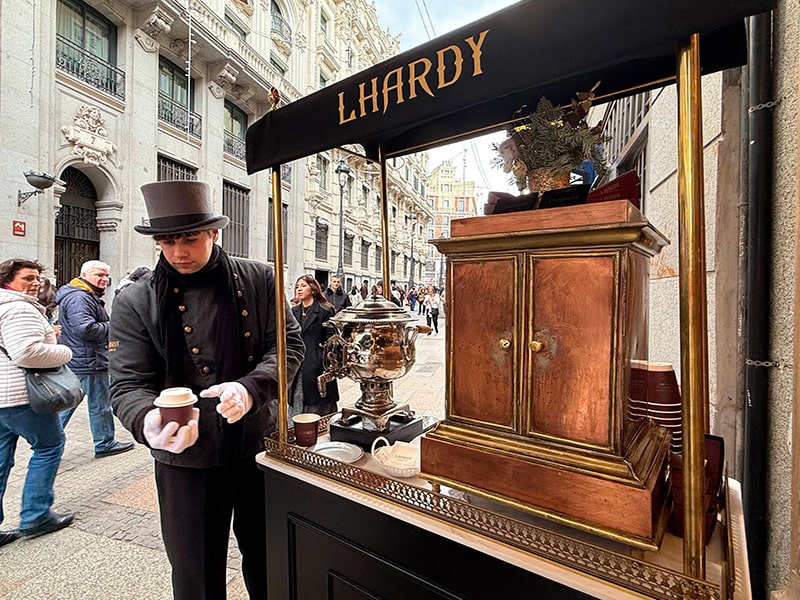 Уличный киоск ресторана Lhardy в Мадриде: сотрудник в классической униформе разливает горячий бульон из самовара, на фоне — историческая улица и прохожие.