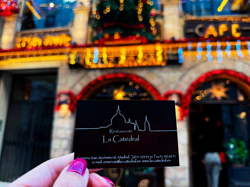 Визитка ресторана La Catedral в руке на фоне рождественской иллюминации фасада кафе в центре Мадрида