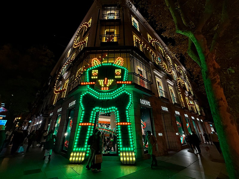 Рождественская иллюминация на фасаде магазина LOEWE на Gran Vía в Мадриде, светящийся портал в виде зеленого кота у входа, огни по всему зданию и прохожие вечером