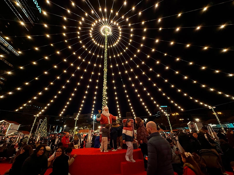 Центр рождественского рынка у El Corte Inglés Castellana в Мадриде: фигура Санта-Клауса и дети под куполом из светящихся гирлянд