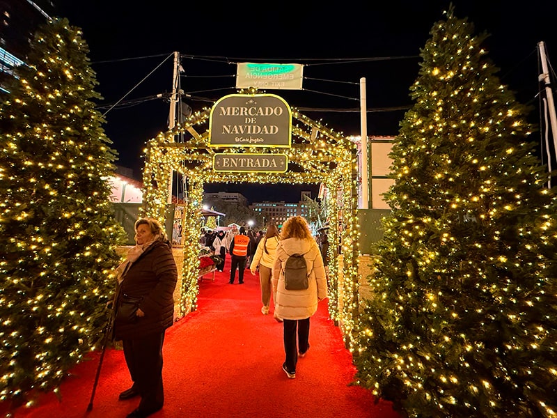Вход на Mercado de Navidad El Corte Inglés в Мадриде: арка с гирляндами, красная ковровая дорожка и люди, входящие на рождественский рынок