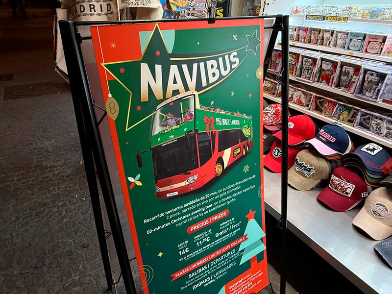 Рекламный стенд рождественского туристического автобуса Navibus в сувенирном киоске в центре Мадрида, рядом витрина с кепками и сувенирами