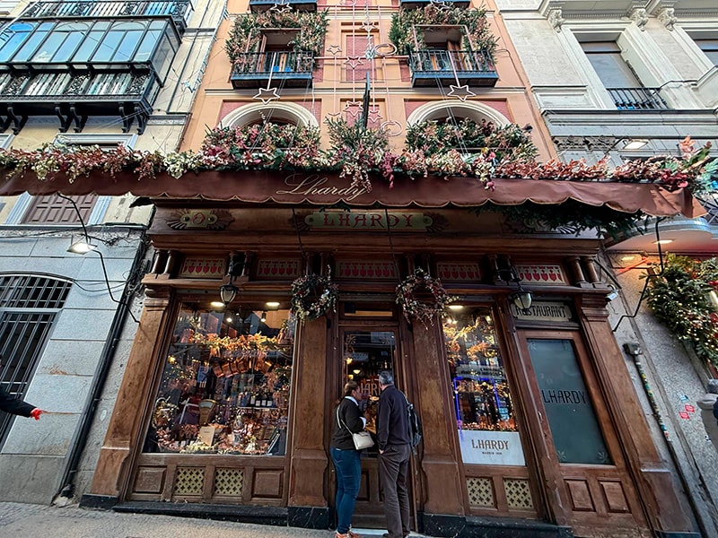 Исторический ресторан Lhardy в Мадриде, фасад украшен рождественскими гирляндами и цветами, люди заглядывают внутрь витрины