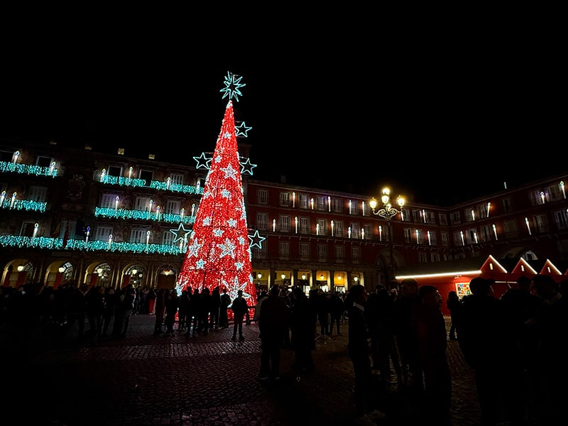 Красная рождественская елка со звездами на Plaza Mayor в Мадриде, вокруг люди гуляют по площади с огнями и домиками рождественского рынка