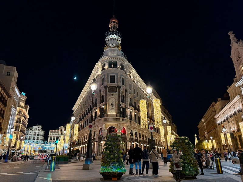 Рождественская иллюминация на Plaza de Canalejas в Мадриде, историческое здание на пересечении Calle de Alcalá с подсветкой, светящиеся гирлянды над улицей и люди, гуляющие вечером