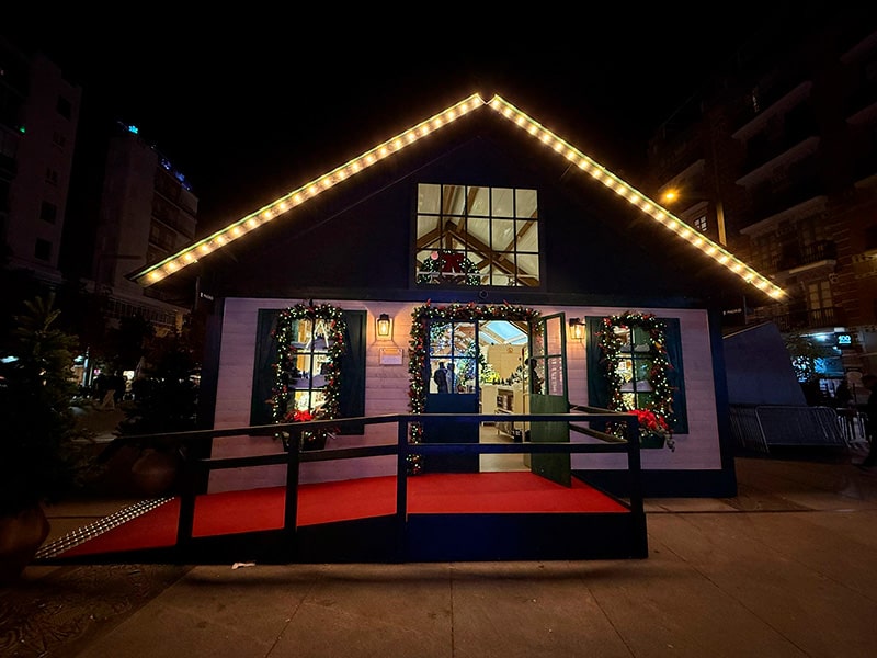 Рождественский домик в Мадриде на городской площади ночью, фасад украшен гирляндами и венками, внутри виден теплый свет и новогодний декор