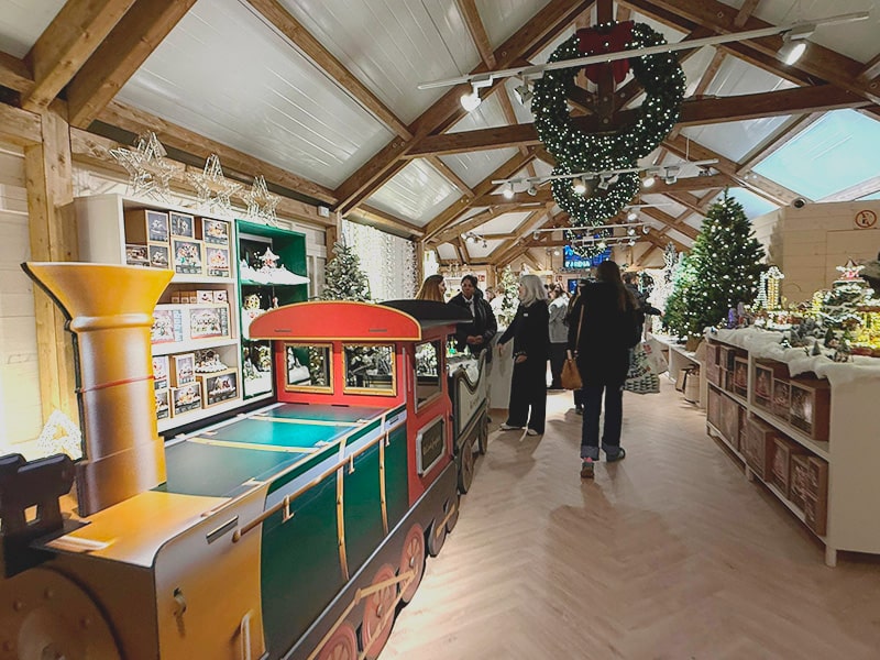 Интерьер рождественского домика: декоративный паровоз, елки, гирлянды и люди, рассматривающие новогодний декор