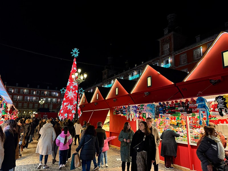 Рождественский рынок на Plaza Mayor в Мадриде, люди гуляют между красными домиками с игрушками и сувенирами, на заднем плане красная елка со звездами