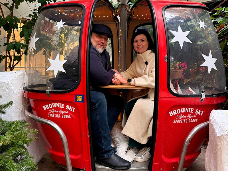 Пара сидит за столиком в красной кабинке-«гондоле» во дворе магазина Brownie в районе Salamanca, Мадрид — уютная зимняя атмосфера и декор со звёздами.