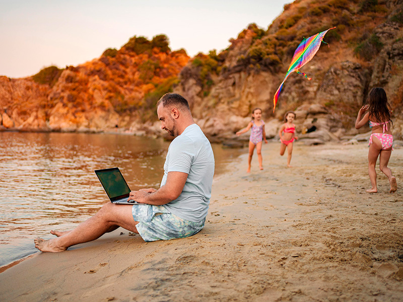 Цифровой кочевник работает с ноутбуком на пляже, пока его дети играют