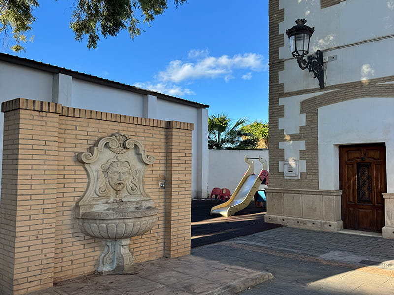Кирпичная стена с каменным фонтаном-барельефом рядом с фасадом здания и детской площадкой с горкой в районе Pobles del Nord, Валенсия.