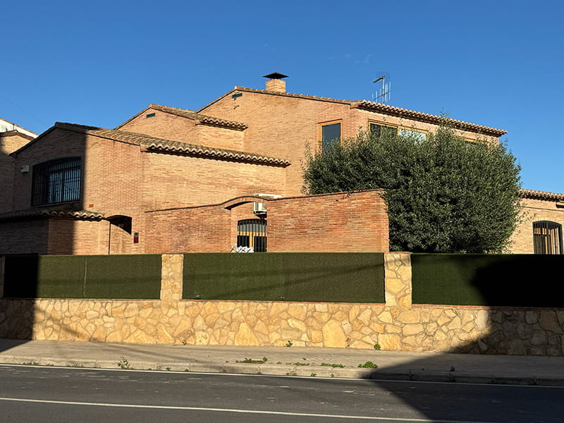 Кирпичный частный дом в Pobles del Nord за каменным забором с зелёной оградой; у фасада растёт большое дерево, солнечный вечерний свет и ясное небо.