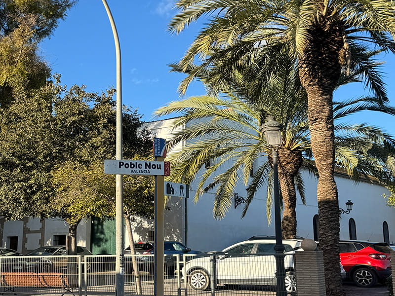 Дорожный указатель «Poble Nou, Valencia» на фоне белого здания, пальм и припаркованных машин; солнечный день, голубое небо.