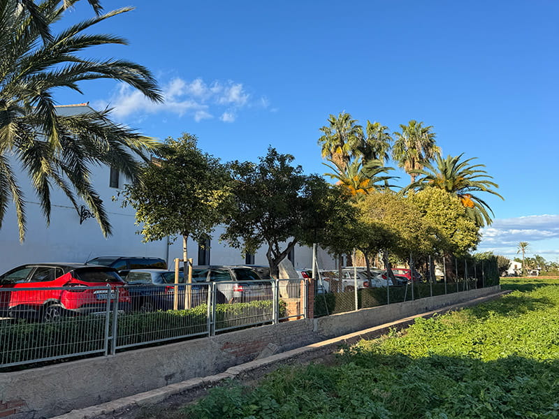 Вид на поля валенсийской уэрты рядом с ограждённой парковкой, белым зданием и пальмами в районе Pobles del Nord, Валенсия.