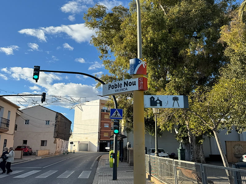 Указатель «Poble Nou, Valencia» у перекрёстка с пешеходным переходом и светофорами, деревенская улица и большие деревья в районе Pobles del Nord.