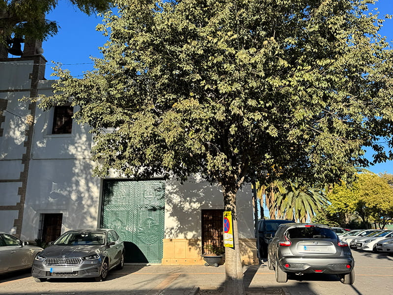 Белое здание с большой зелёной дверью и парковкой у фасада, перед ним — крупное дерево и припаркованные машины в Poblados del Norte (Валенсия).