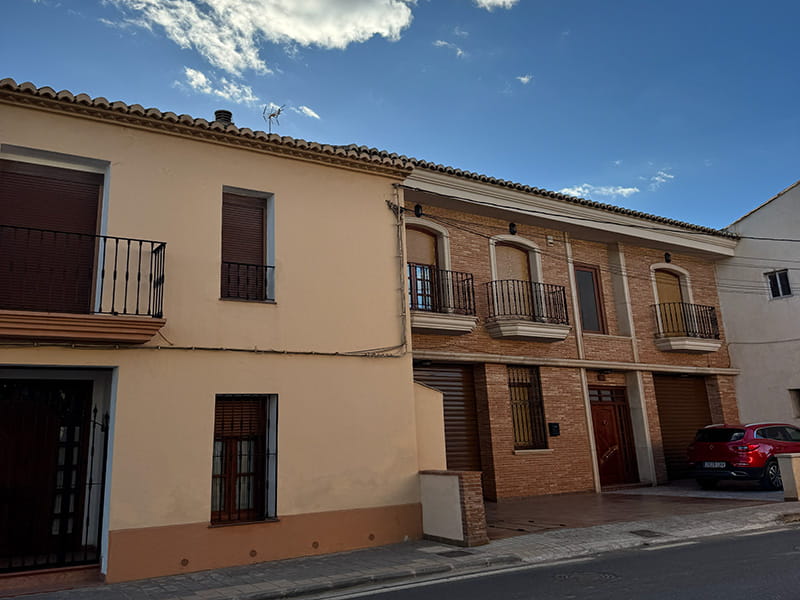 Улица в Poblados del Norte (Валенсия): двухэтажные сельские дома с балконами и ставнями, кирпичный фасад и припаркованный автомобиль.