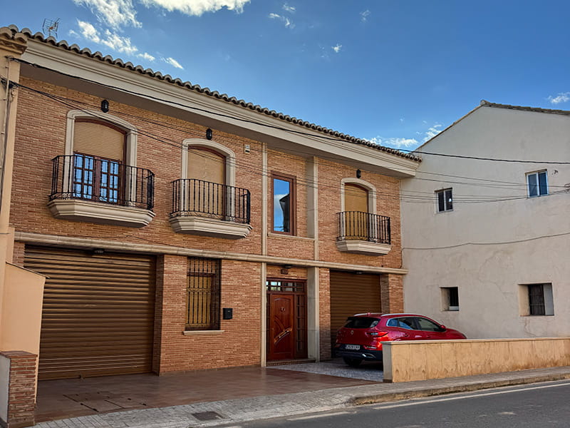 Двухэтажный кирпичный дом с тремя балконами и ставнями на тихой улице Poblados del Norte; у фасада припаркован красный автомобиль, ясное небо.