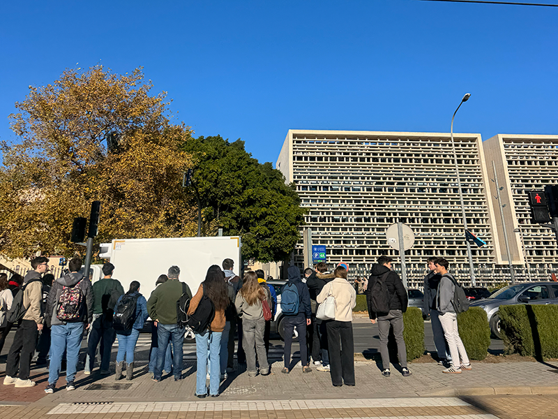 Группа студентов ждёт транспорт на перекрёстке в районе Algirós, Валенсия: люди с рюкзаками стоят у дороги на фоне современных зданий и деревьев.