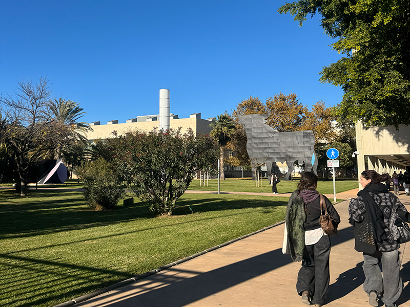 Студенты идут по дорожке через зелёный кампус в районе Algirós (Валенсия): газоны, деревья и современные корпуса университета на фоне ясного неба.