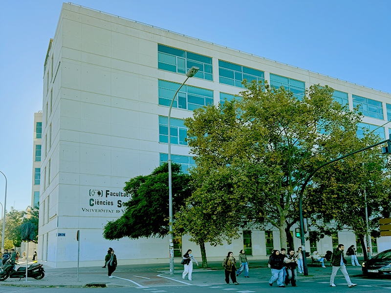 Белое здание факультета Universitat de València в районе Algirós: современный корпус с голубыми окнами, деревья и студенты на улице.