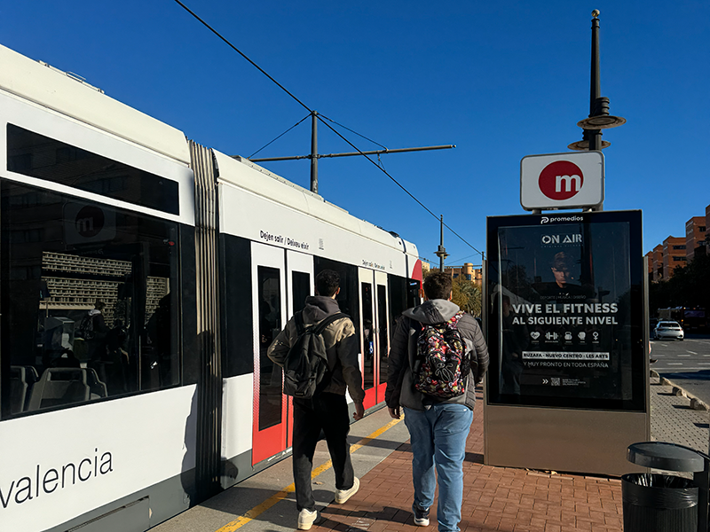 Остановка Metrovalencia в районе Algirós (Валенсия): двое людей с рюкзаками идут вдоль платформы рядом с бело-красным поездом, рядом знак метро и рекламный стенд.
