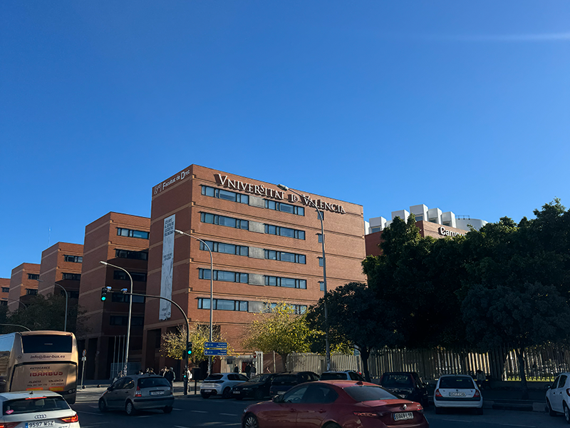 Здание Universitat de València в районе Algirós, Валенсия: кирпичный корпус университета на фоне ясного голубого неба, перед ним городская улица с автомобилями.