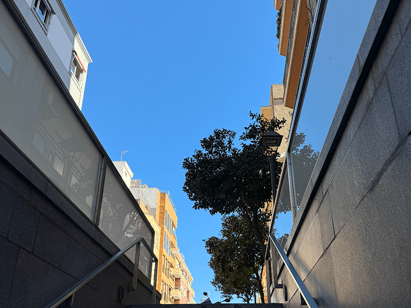 Выход из подземного перехода у станции Benimaclet в Валенсии: лестница с перилами, деревья и жилые дома на фоне голубого неба.