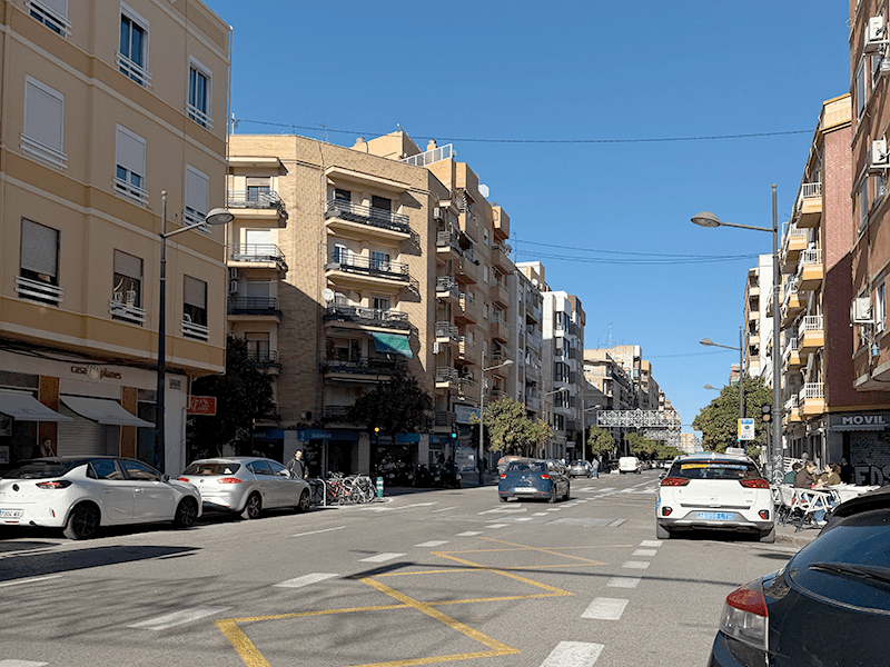 Широкая улица в районе Benimaclet, Валенсия: жилые дома с балконами, припаркованные машины и ясное голубое небо.