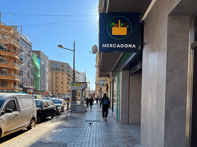 Улица в районе Benimaclet, Валенсия: вывеска супермаркета Mercadona на фасаде и пешеходная зона с припаркованными машинами.