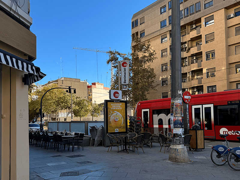 Станция метро Benimaclet в Валенсии: красный трамвай Metrovalencia у остановки и терраса кафе на площади.
