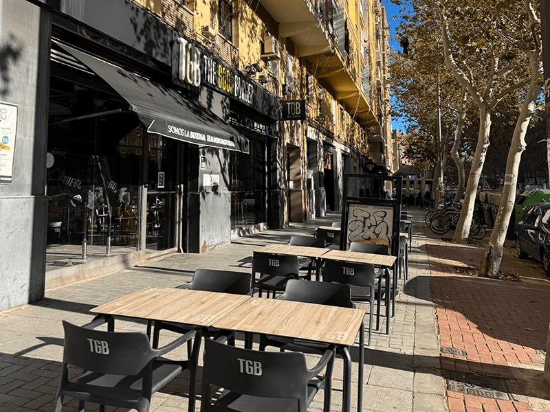 Терраса кафе в районе Benimaclet, Валенсия: столики на тротуаре под навесом и аллея с платанами вдоль улицы.